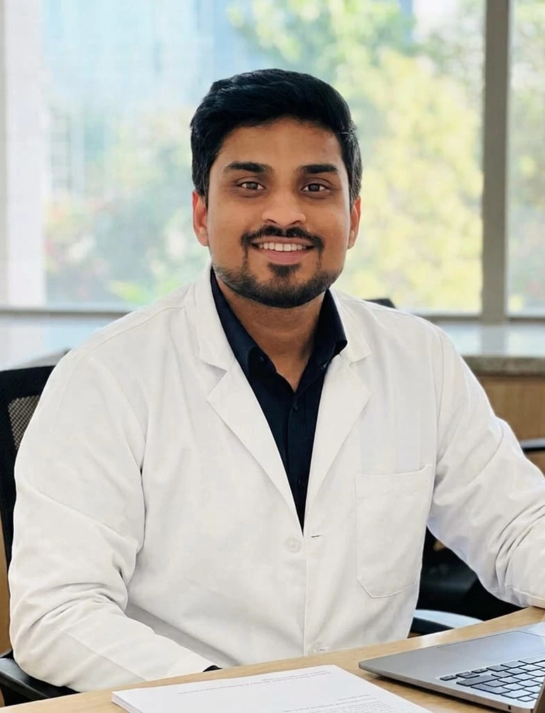 5 | Neo Dental | Neo Dental Smiling male doctor in a white coat seated at a desk with a laptop in front of him.