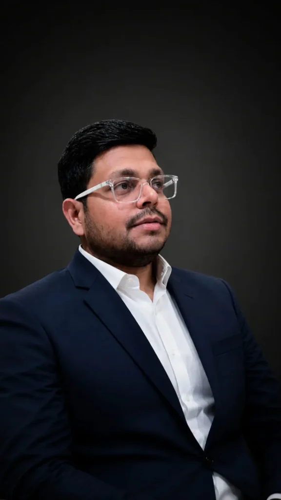 Professional headshot of a man with dark hair and clear glasses, wearing a navy suit and white shirt against a dark backdrop.